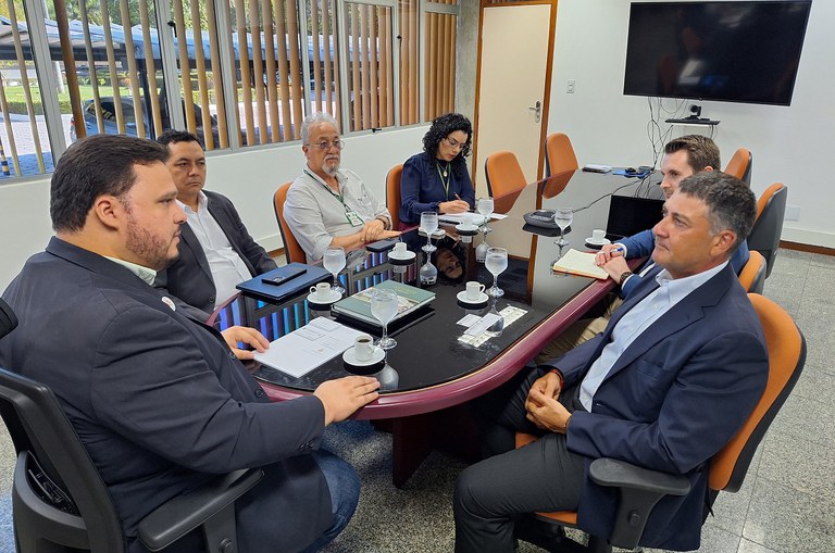 Foto mostra uma reunião institucional com seis pessoas numa sala, entre elas uma mulher, que está anotando os principais pontos da conversa