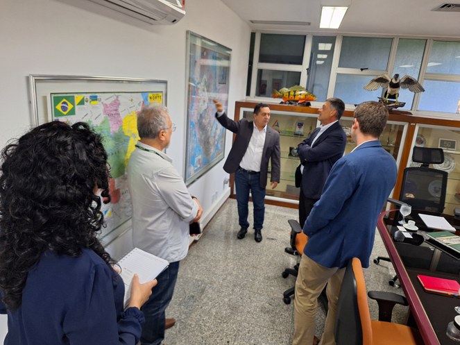 Foto mostra senhor apontando para um mapa na parede, com o intuito de explicar uma situação a visitantes de uma reunião