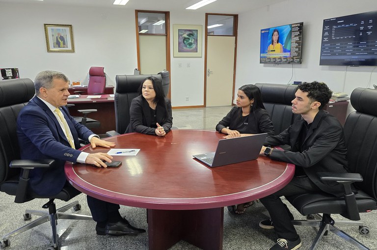 Dois homens e duas mulheres sentados ao redor de uma mesa redonda. As mulheres e um homem jovem estão de rou´pa preta e o outro homem de meia idade, de terno