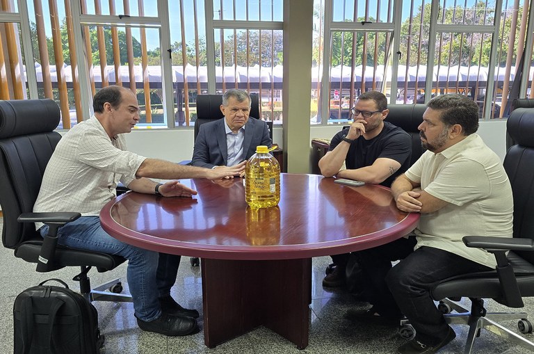 Imagem mostra quatro homens heteros brancos de meia idade ao redor de uma mesa redonda, ao centro da mesa uma garrafa de óleo de cinco litros.