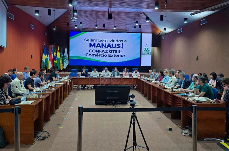 Foto mostra homem de camisa branca falando à mesa, numa reunião com outras pessoas e com um telão de apoio ao fundo