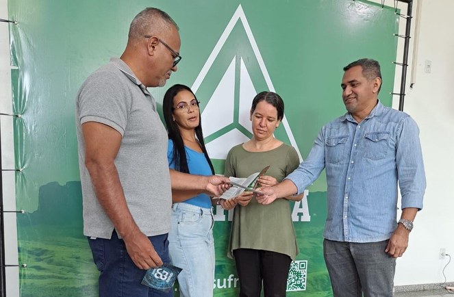 Foto mostra pessoas olhando para folders, com banner ao fundo