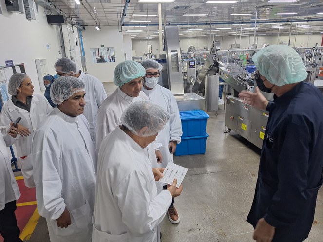 Foto mostra homem de touca e bata brancas observando um objeto, acompanhado de outras pessoas