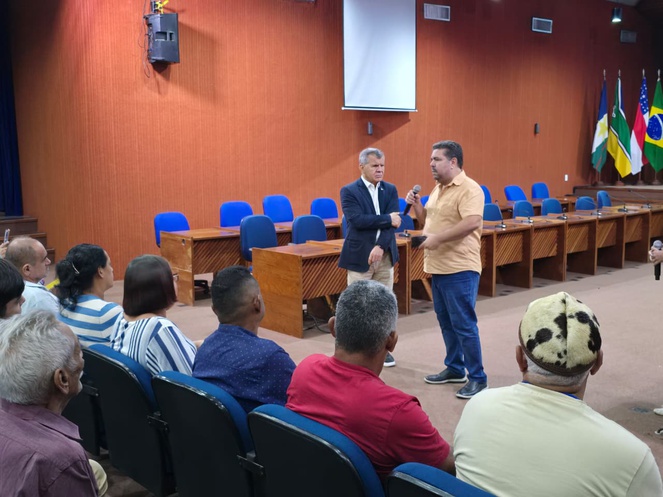 Foto mostra homem de blusa bege falando em um evento dentro de um auditório, ao lado de um homem vestido de paletó