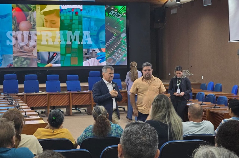 Foto mostra homem de paletó falando em evento ao lado de outro homem