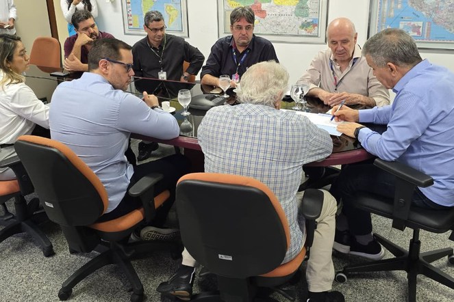 Foto mostra homem escrevendo num papel, observado por grupo de pessoas