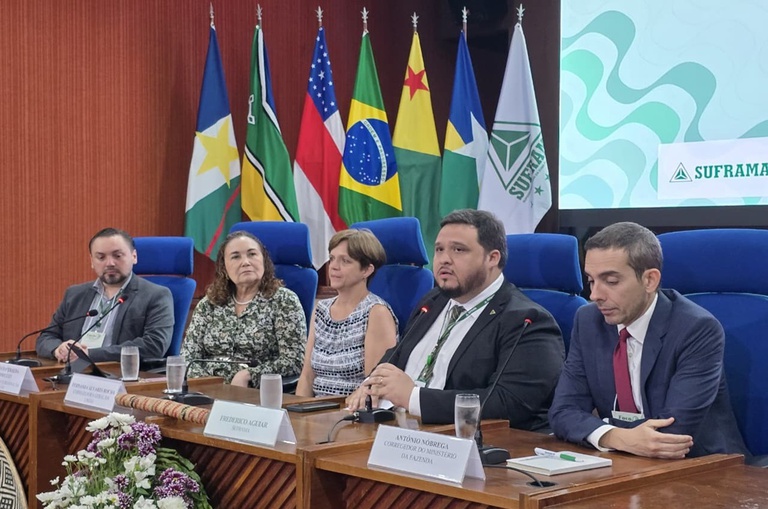 Foto mostra homem de blazer preto falando ao microfone ao lado de outras pessas à mesa