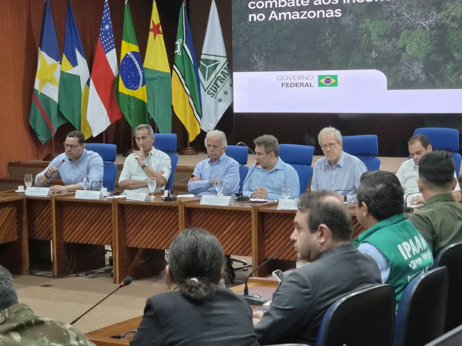 Foto mostra homem falando ao microfone ao lado de outros homens e um telão ao fundo
