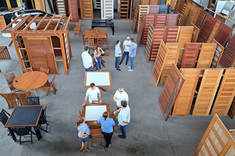 Foto mostra homem de óculos escuro conversando com pessoas numa loja de móveis em madeira
