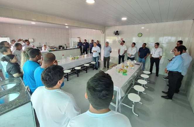 Foto mostra homem de blusa branca conversando com grupo de pessoas em um refeitório