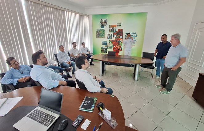 Foto mostra homem em pé de blusa azul clara fazendo uma explanação a grupo de pessoas