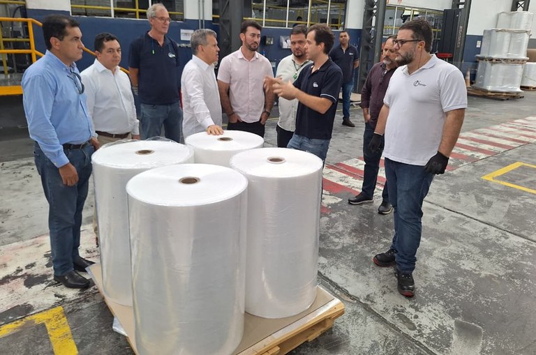 Foto mostra homem de blusa azul escuro falando com outro homens numa fábrica de plásticos