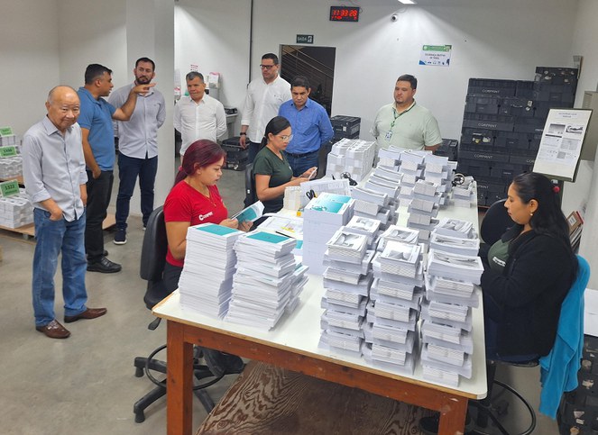 Foto mostra mulheres manuseando blocos de papel, observadas por um grupo de homens