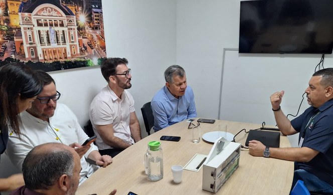 Foto mostra homem de blusa azul fazendo uma explanação a grupo de pessoas com um telão ao fundo