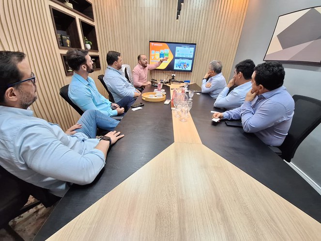 Foto mostra pessoas sentadas à mesa olhando para um telão