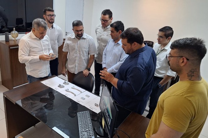 Foto mostra um grupo de pessoas observando peças para fabricação de automóveis que estão em cima de uma mesa