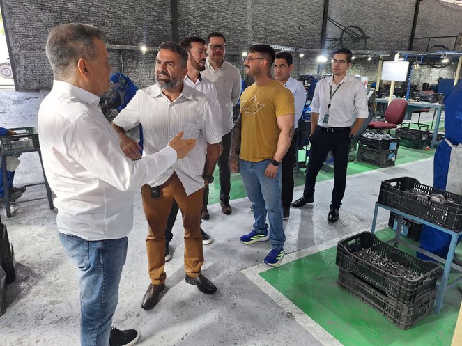 Foto mostra homem de blusa branca falando a outro homem, acompanhado de outras pessoas e peças para fabricação de automóveisde automóveis que estão em cima de uma mesa