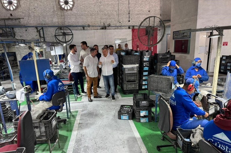 Foto mostra grupo de pessoas observando peças para fabricação de automóveis que estão em cima de uma mesa