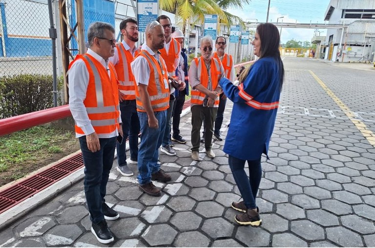 Foto mostra mulher de jaleco azul fazendo uma explanação a grupo de pessoas