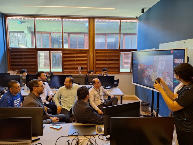 Foto mostra uma mulher mostrando imagens num telão, observada por grupo de pessoas