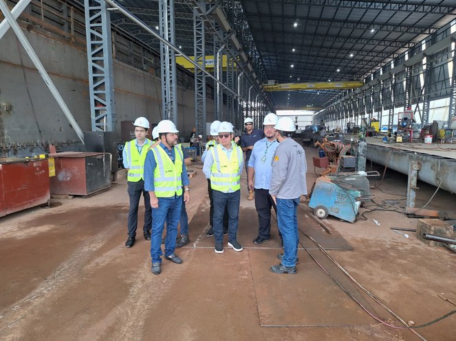 Foto mostra grupo de pessoas com capacete e jaquetas de proteção dentro de um estaleiro