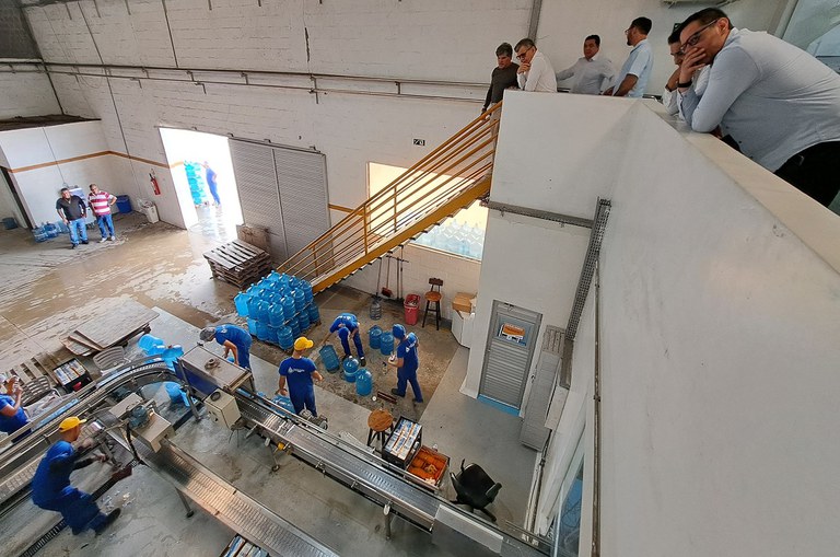 Foto mostra pessoas olhando do alto, outras pessoas em baixo trabalhando na produção de garrafas de água mineral 