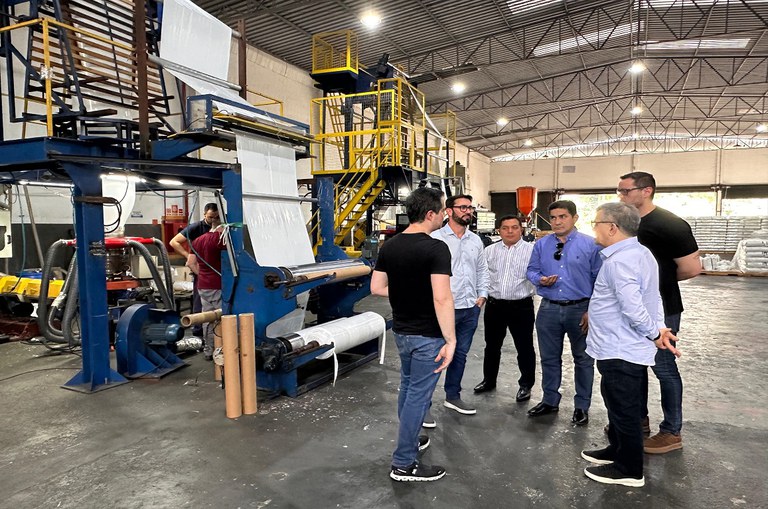 Grupo de pessoas, em sua maioria homens em um galpão de fábrica, próximo à uma maquina