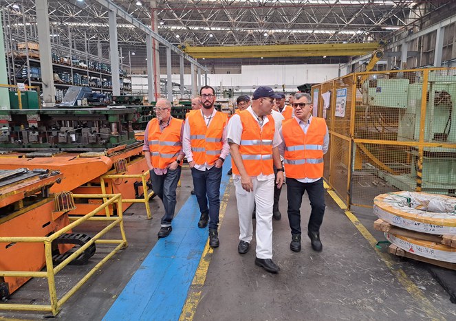 Foto mostra homens andando em uma fábrica, conferindo uma linha de produção de peças para motocicletas