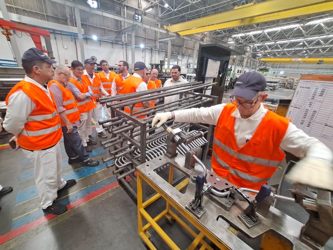 Foto mostra homem de boné manuseando um equipamento em linha de produção de fábrica, observado por outros homens
