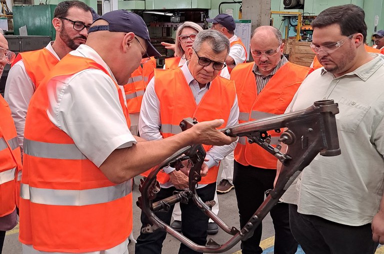 Foto mostra homem de boné mostra uma peça de motocicleta a um grupo de visitantes em uma fábrica