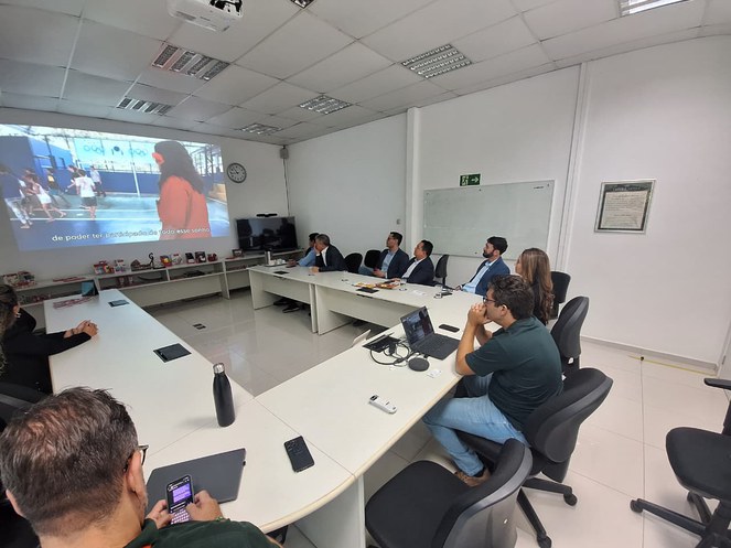 Foto mostra grupo de pessoas sentadas e assistindo a uma apresentação no telão