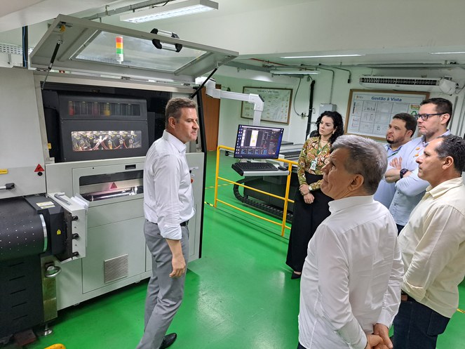 Foto mostra homem de blusa branca mostrando uma máquina de fabricar papelão a um grupo de visitantes em uma fábrica