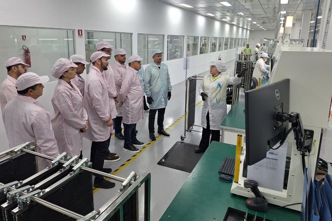 Foto mostra mulher de bata branca mostrando equipamento a um grupo de pessoas também de bata