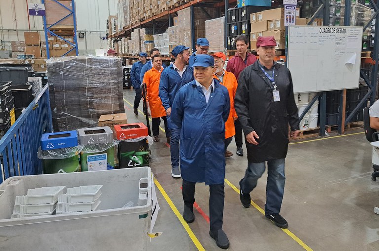 Foto mostra dois homens de boné e jaleco caminhando numa fábrica, seguidos de outros homens