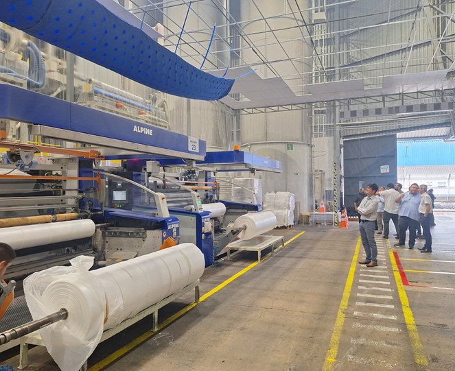 Foto mostra homem de braços cruzados, acompanhado de outros homens, observando uma linha de produção de produtos plásticos