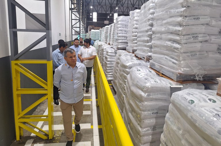 Foto mostra homem vestido com blusa azul claro e calça bege, observando um aglomerado de sacos, com outros homens atrás dele