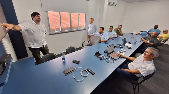 Foto mostra homem de blusa branca e calça verde, explanando para um grupo de outros homens em uma sala fechada