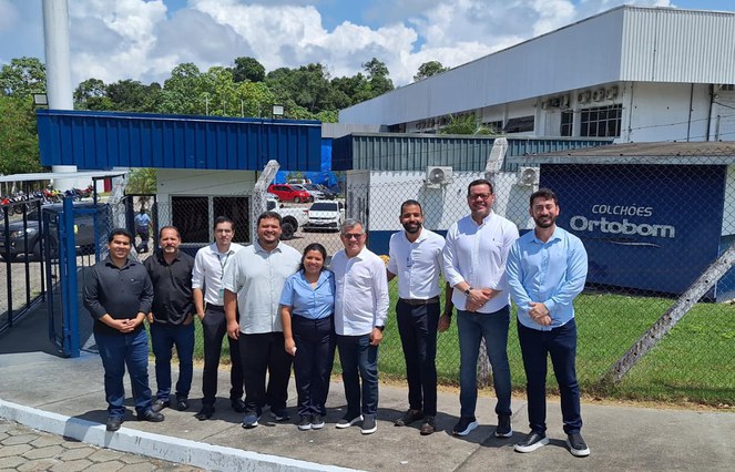 Foto mostra pessoas posando para foto tendo fachada de uma fábrica ao fundo