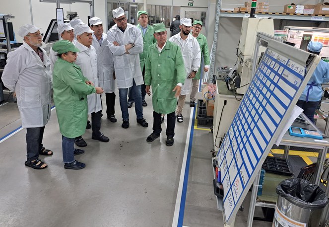 Foto mostra homem de jaleco verde mostrando um quadro explicativo a um grupo de pessoas em visita a uma fábrica