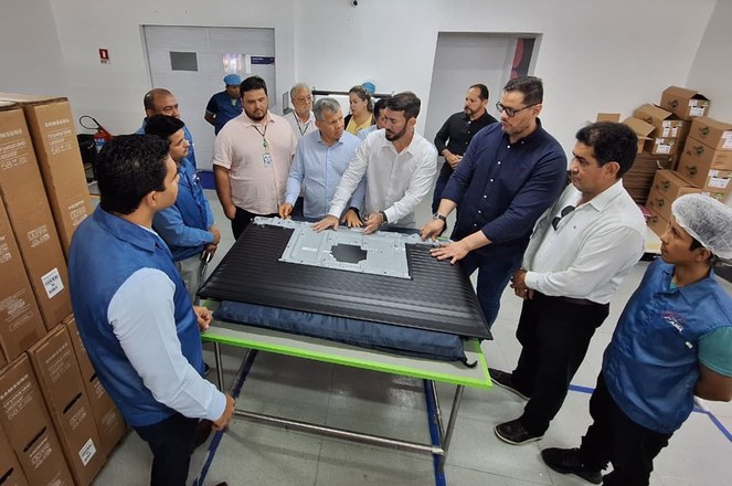 Foto mostra homem de blusa branca com a mão em um objeto, observado por grupo de pessoas