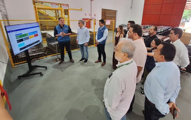 Foto mostra homem com jaqueta azul fazendo uma explanação a grupo de pessoas, com telão como apoio