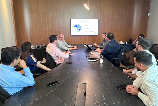 Foto mostra pessoas numa reunião, sentadas à mesa e com o apoio de telão