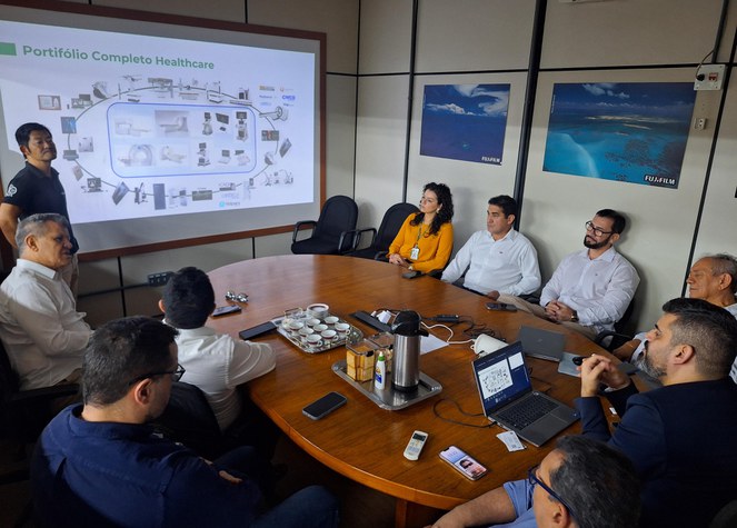Foto mostra homem de blusa branco falando com outros homens observando, e um telão ao fundo