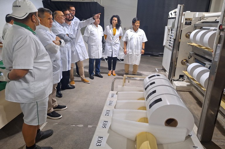 Foto mostra homens vestidos de jaleco branco observando uma máquina com papel