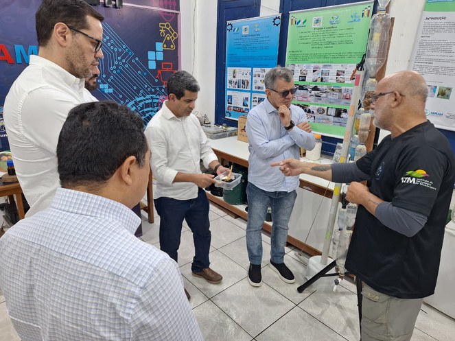 Foto mostra homem de blusa escura apontando para objeto que outro homem tem nas mãos