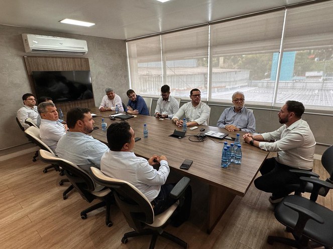 Foto mostra pessoas à mesa, numa reunião dentro de uma sala com janelas de vidros transparentes
