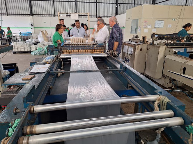 Foto mostra um homem de blusa branca mexendo em produto plástico, observado por um grupo de pessoas