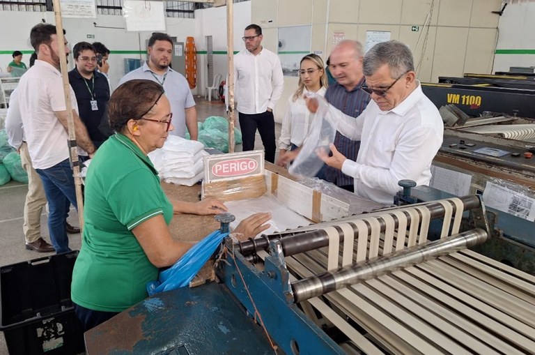 Foto mostra homem de blusa branca mexendo em produto plástico, observado por um grupo de pessoas 