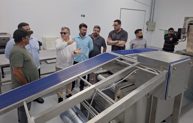 Foto mostra homem de óculos escuros falando a outros homens em frente de uma máquina