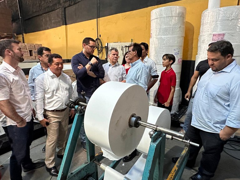 Imagem mostra pessoas em pe vendo uma linha de produção de papel para guardanapos